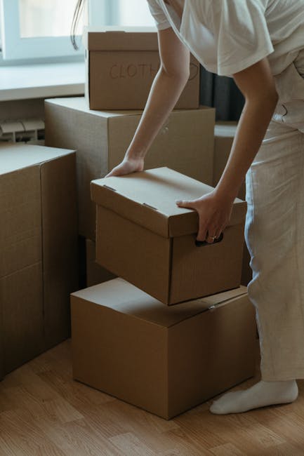 A person wearing white clothing and standing on a wooden floor is in the process of packing or unpacking moving boxes made of brown cardboard, with one box labeled 'CLOTHES' visible in the background. The individual is holding a smaller box, carefully placing it on a stack of three boxes, with other stacked boxes surrounding them. The scene is indoors, near a window that provides natural light, and furniture or additional boxes may be present in the background. This image illustrates the packing stage of a home relocation, highlighting the use of sturdy cardboard boxes and proper handling during furniture transport, as part of a professional removals process by Man and Van New Malden.