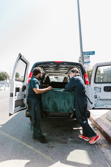 Two movers, wearing navy uniforms and protective eyewear, are loading a large, dark green upholstered piece of furniture into the back of a black moving van parked on a street. The van's rear doors ar