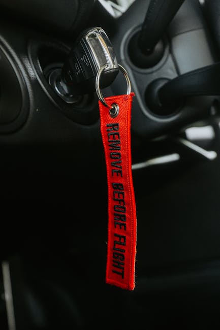 Close-up of a car key inserted into the ignition switch inside a vehicle, with a red fabric keychain attached, displaying the words 'REMOVE BEFORE FLIGHT' in black text. The view captures part of the vehicle's dashboard and steering column, with visible black plastic and grey interior materials. The ignition area is slightly illuminated by ambient interior lighting. This image relates to home relocation and moving services provided by Man and Van New Malden, showcasing the transportation aspect of furniture transport and packing during house removals, emphasizing readiness for a home relocation or moving process.