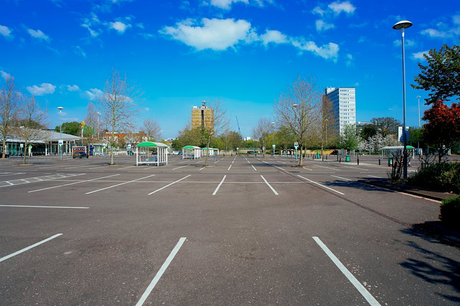 A wide view of an empty parking lot in New Malden under a bright blue sky with scattered clouds, featuring clearly marked white parking spaces and a few scattered trees with budding leaves. In the background, there are modern multi-storey office buildings and smaller structures, with some covered bus stops or kiosks with green and white umbrellas near the edges of the lot. The area is well-lit with tall street lamps and has a few vehicles parked along the perimeter. The foreground shows the beginning of a loading zone, where a professional moving team from Man and Van New Malden is executing a home relocation, carefully lifting and carrying furniture wrapped in plastic and blankets, with a trolley and straps visible for securing items inside a large van. The scene captures the organized loading process typical during house removals, complete with packing materials like cardboard boxes and furniture pads, aligned with local Kingston Council parking rules for New Malden removals.