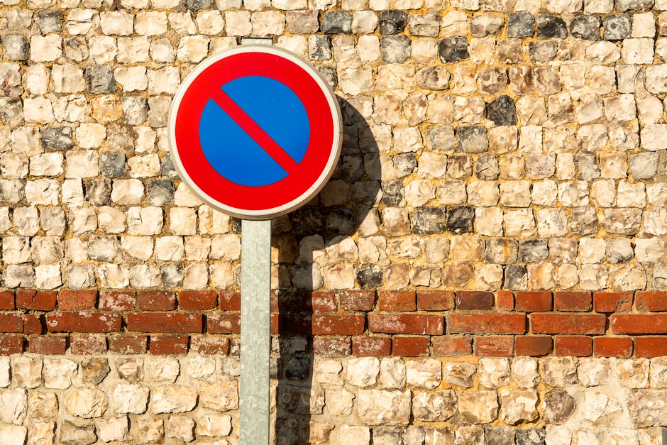 A circular traffic restriction sign with a red border, blue background, and a red diagonal line across the center, mounted on a gray metal pole. The sign is set against a textured stone wall composed of beige and brown stones with a horizontal band of red bricks at the lower portion. The shadow of the sign extends onto the wall, indicating sunlight. This scene is relevant to house removals and logistics, as parking restrictions impact loading and unloading areas during home relocation and furniture transport processes, which companies like Man and Van New Malden manage carefully to comply with local parking rules in Kingston and New Malden.