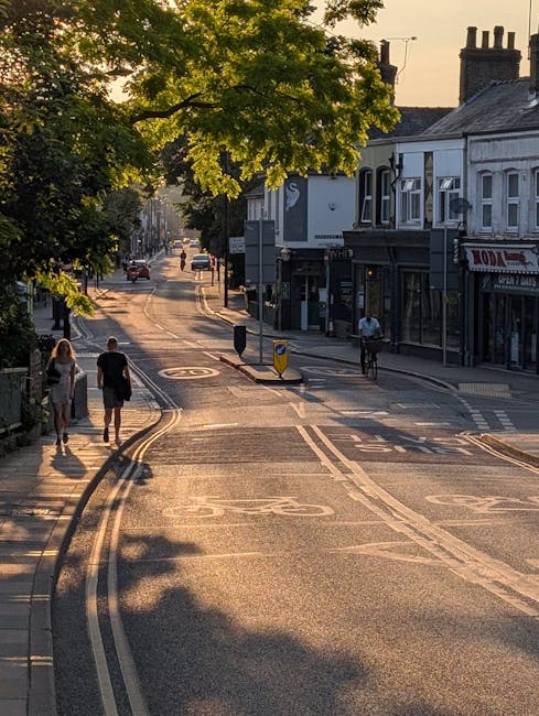 A high-angle view of Kingston Road in New Malden showing a narrow, one-lane road with parked cars along both sides and moving vehicles in both directions, including a red double-decker bus and various