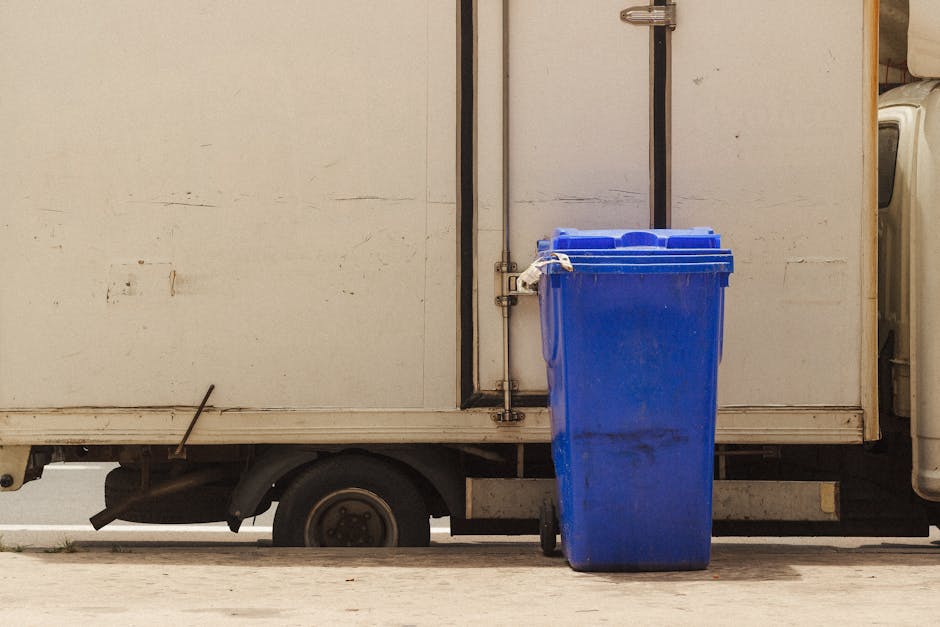 Photograph from outside a residential property showing a white delivery van with a closed side door parked on the street. In front of the van, a large blue plastic recycling bin with a closed lid is p