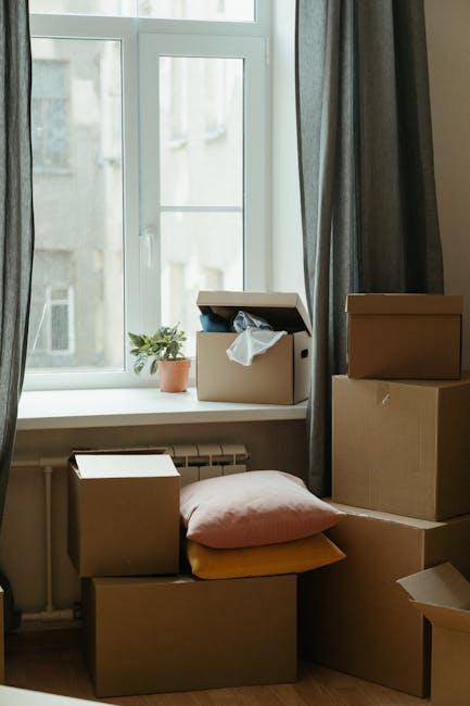A collection of cardboard moving boxes, some with flaps open revealing packing materials such as plastic wrapping and fabric, are stacked on a wooden floor inside a room near a large double-glazed window with white frames. The boxes vary in size and are placed both directly on the floor and on each other. A pink cushion and a yellow pillow are positioned on top of some of the boxes, indicating possible use as seating or support during packing or loading processes. The window sill features a small potted plant with green foliage, adding a natural element to the scene. Outside the window, blurred views of neighboring buildings are visible, suggesting an urban residential environment. Dark curtains are drawn back to allow natural light to illuminate the space. This image exemplifies typical household packing preparations that are part of a home relocation process managed by [COMPANY_NAME], specializing in removals and furniture transport services, including packing and loading activities for moves from flats in the KT3 postcode area in New Malden.
