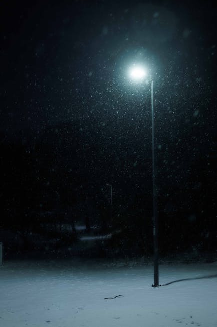 A tall street lamp illuminated from below, casting a bright light on the snow-covered ground during a snowy night. Snowflakes are visibly falling, creating a layered white scene, and the dark sky and background are faintly visible. The lamp is positioned near the edge of a pavement or driveway outside a residential area, with no other objects or vehicles in view. The scene captures the quiet atmosphere typical of winter evenings, with the lighting and snow emphasizing a peaceful, cold environment, relevant to home relocation or transport logistics as part of the moving process discussed on the New Malden Station move checklist page, with Man and Van New Malden providing professional removals services.