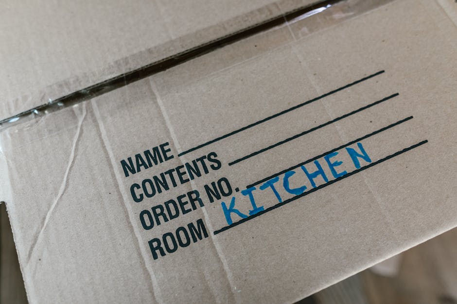 Close-up of a cardboard moving box with printed fields for name, contents, order number, and room. The room field is handwritten in blue ink as 'kitchen.' The box is sealed with clear packing tape and appears to be part of a home relocation or packing process, with the box placed among other moving supplies. The background is blurred, indicating an indoor environment, likely within a residential property or during the loading process of furniture transport. This image reflects organized packing activities associated with a professional removals service, such as those offered by Man and Van New Malden, supporting smooth house removals and furniture transport during a home move.