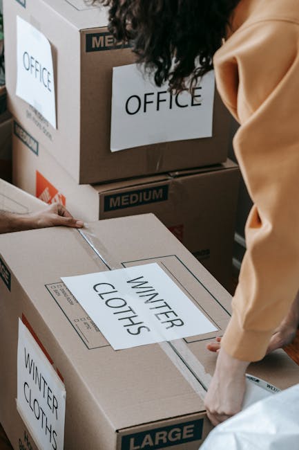 Close-up view of a professional removals process inside a property, showing several medium and large cardboard boxes labeled with signs such as 'OFFICE' and 'WINTER CLOTHES' being handled by a mover wearing a beige shirt. The boxes are secured with packing tape and are positioned on a floor near a doorway or entrance. One person is seen gripping the top of a large box, preparing for transport or loading onto a vehicle, while other boxes are stacked against a wall. The environment appears well-lit, indicating a daytime setting, and the scene is part of a house or office relocation where packing and furniture transport are being organized, with furniture or other items possibly being prepared for movement as observed in the detailed arrangement and careful handling by Man and Van New Malden.