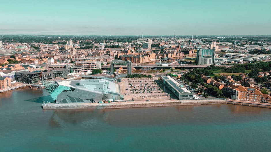 An aerial view of a riverside urban area showing a modern commercial or office building with a glass facade located on the waterfront, with adjacent parking lots filled with vehicles. In the foreground, a street with pedestrians is visible, while behind the building, there are densely packed residential and commercial structures with brick facades and rooftops. The background features a cityscape with various high-rise and mid-rise buildings, bridges crossing the river, and a clear sky overhead. The scene captures the environment where home relocation services by Man and Van New Malden may operate, illustrating the typical setting for furniture transport and moving logistics in the Royal Borough of Kingston upon Thames.