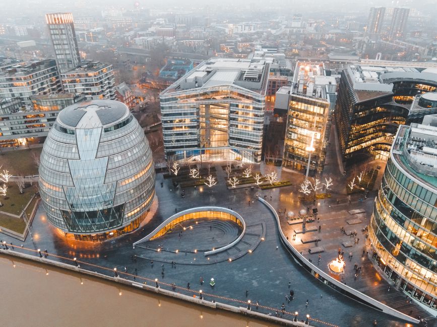 An aerial view of a modern cityscape in the evening, featuring several contemporary office buildings with glass facades illuminated from within. In the foreground, there is a paved public square with a circular water feature, surrounded by white leafless trees adorned with string lights. The area includes benches and small sculptures, with pedestrian pathways and a few people walking. To the left, a distinctive rounded building with a patterned glass exterior is visible. Surrounding the square are additional office blocks and commercial structures, with some under construction or renovation, set against a distant hazy skyline of high-rise buildings. The lighting from the buildings, streetlights, and decorative elements enhances visibility, highlighting the urban environment where commercial property management and urban planning activities related to city development and relocation services could take place. This scene exemplifies the type of city infrastructure associated with professional moving and furniture transport during urban home relocation projects.