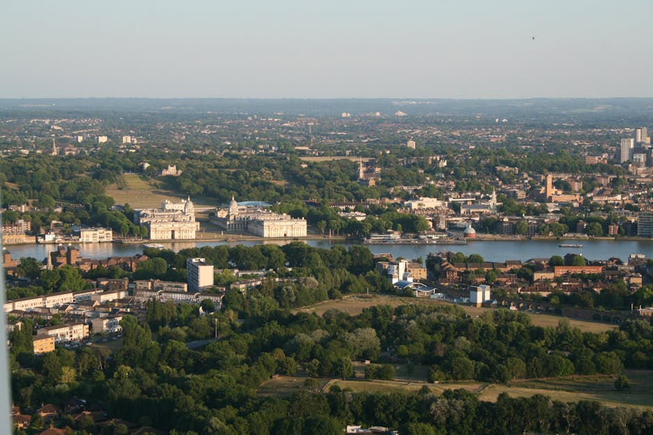 An aerial view of a riverside urban area showing a modern commercial or office building with a glass facade located on the waterfront, with adjacent parking lots filled with vehicles. In the foreground, a street with pedestrians is visible, while behind the building, there are densely packed residential and commercial structures with brick facades and rooftops. The background features a cityscape with various high-rise and mid-rise buildings, bridges crossing the river, and a clear sky overhead. The scene captures the environment where home relocation services by Man and Van New Malden may operate, illustrating the typical setting for furniture transport and moving logistics in the Royal Borough of Kingston upon Thames.
