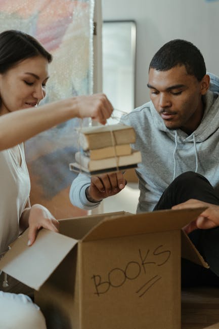 Two individuals engaged in packing activities during a house move; the woman on the left, with dark hair, is placing items into a cardboard box labeled 'BOOKS,' while the man on the right, with short dark hair, is holding a small stack of books wrapped together with packing materials. They are seated on the floor inside a home, near a doorway or open space, with a cardboard box in front of them. The box appears to be partially filled, with additional packing supplies such as plastic wrap or paper nearby. The background features a blurred interior setting, suggesting the move is in progress. This scene illustrates the packing process involved in home relocation, managed by professional removals services such as Man and Van New Malden, focused on careful handling of personal belongings during a furniture transport or moving operation.