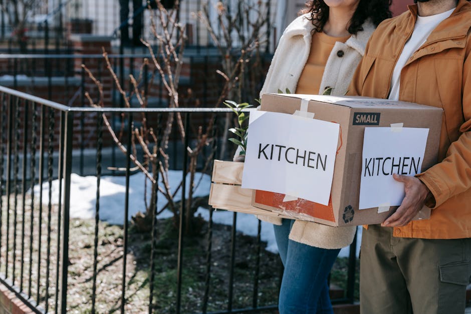 A person holding a cardboard box labeled 'KITCHEN' with a smaller wooden crate and another box secured with tape, as they prepare to load residential items during a house relocation. The scene is set outdoors on a paved area next to a black metal fence, with some snow on the ground and leafless bushes in the background, indicating winter weather. The individual is wearing casual clothing, including a beige sweater and a tan jacket, while standing beside another person. This image illustrates packing and moving processes typical of home removals services, such as those offered by Man and Van New Malden, emphasizing careful handling of kitchen items and organized packing for efficient furniture transport and home relocation.