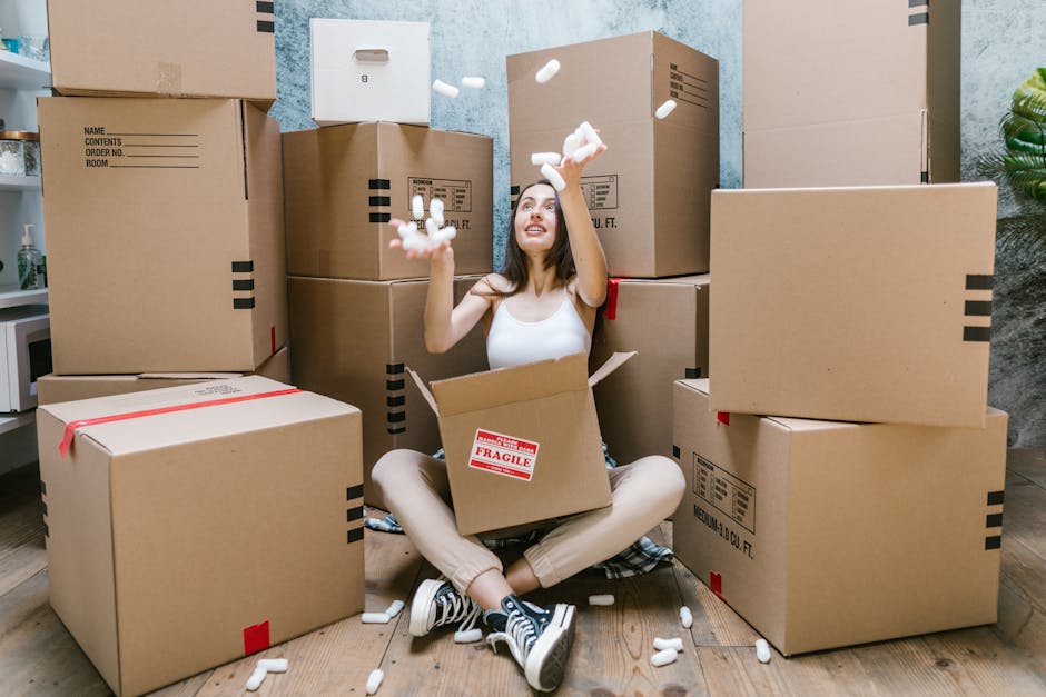 A young woman with long dark hair, wearing a white tank top, beige pants, and black sneakers with white laces, is sitting cross-legged on a wooden floor inside a room. She is surrounded by multiple large cardboard boxes, some sealed with red tape, placed against the walls and on the floor. She is inside an open box, holding handfuls of white packing foam or packing peanuts, which are also scattered around her and in mid-air, as she playsfully throws them upward. The boxes vary in size, with some labeled for contents or weight, and are positioned behind and around her, suggesting she is in the process of packing or unpacking during a home relocation. The scene is well-lit with natural or soft lighting, and the background features a lightly painted wall with a launching plant visible on the right side, reflecting the typical environment for house removals and furniture transport as part of a professional moving process, associated with Man and Van New Malden.